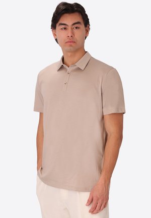 Uomo che indossa una polo beige a maniche corte e pantaloni bianchi, in piedi con le mani nelle tasche, rivolto leggermente a sinistra su uno sfondo uniforme.