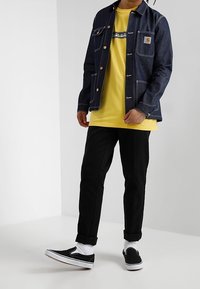 Chaqueta de mezclilla en azul oscuro con botones dorados, camiseta amarilla, pantalones negros y zapatos negros sin cordones con suelas y puños blancos.