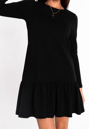 Kobieta w czarnej, długorękawowej sukience z falbaną u dołu i złotym naszyjnikiem z krzyżykiem, stojąca na tle jednolitej białej ściany.