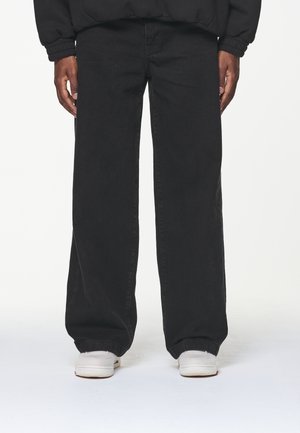 Osoba v pohodlných černých džínách a bílých teniskách stojící s rukama uvolněně podél těla před jednoduchým pozadím.