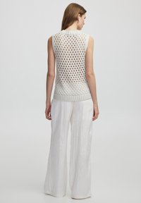 Top sin mangas de punto con un patrón de tejido suelto y abierto en crema, que presenta un escote y dobladillo de canalé. Combinado con pantalones ligeros blancos.