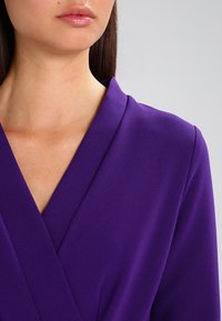 Lilla omslagkjole med dyb V-hals og glat tekstur. Stoffet ser struktureret ud med en diskret glans, der fremhæver dens intense farve.