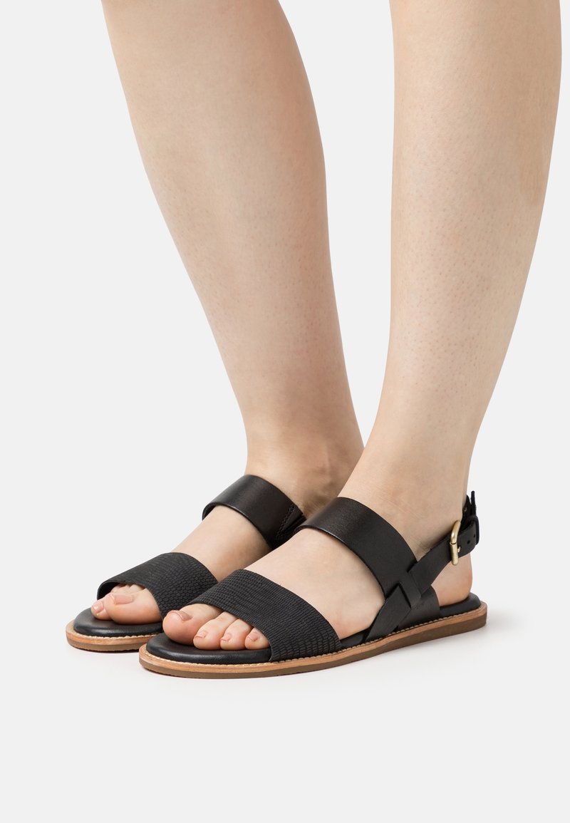 Pieds nus portant des sandales en cuir noir avec de larges brides texturées et des fermetures à boucle à la cheville sur un fond gris clair.