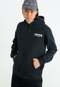 Sudadera negra de algodón con un bolsillo canguro delantero y el logo blanco de "Converse All-Star". El modelo también lleva una gorra gris con acentos metálicos.