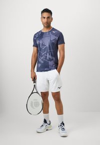 Tricou sportiv cu mânecă scurtă de culoare albastru închis, cu un model geometric, asortat cu pantaloni scurți albi și încălțări de tenis, ținând o rachetă de tenis.