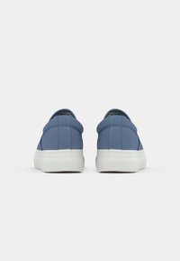 Ballerines en toile bleue avec une semelle en caoutchouc blanc texturé et un col rembourré, présentant un design minimaliste et sans lacets visibles.