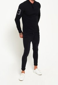 Polo noir à manches longues avec col et patte de boutonnage, arborant le numéro 3 gris sur la manche gauche. Associé à un jean skinny noir et des baskets blanches.