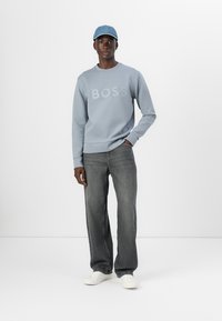 Felpa azzurro chiaro con logo "BOSS", abbinata a jeans grigio scuro e sneakers bianche. Il modello indossa un berretto azzurro chiaro.