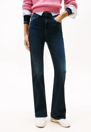 Person iført mørkeblå bootcut jeans, hvide sneakers, en pink strikket sweater over en lagdelt langærmet skjorte med ærmerne rullet op.
