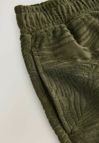 Gros plan d'un short en tissu texturé vert olive avec une ceinture élastique et une poche latérale sur une surface claire.