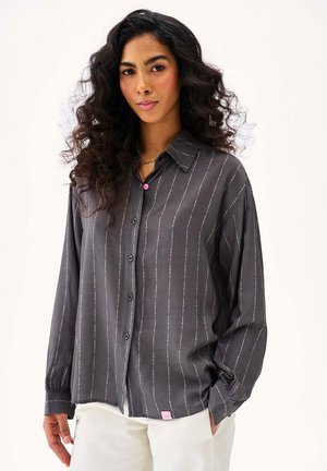 Kobieta o długich kręconych włosach, ubrana w luźną ciemnoszarą koszulę zapinaną na guziki z pionowymi, kropkowanymi paskami oraz białe spodnie, na jasnym tle.
