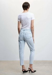Femme debout de dos, portant un t-shirt blanc, un jean taille haute bleu clair, des chaussures à talons ouvertes noires, et un petit tatouage sur le bras.