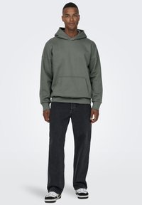 Hooded sweatshirt in olive green cotton blend with a front pocket, paired with black wide-leg jeans and black-and-white sneakers.