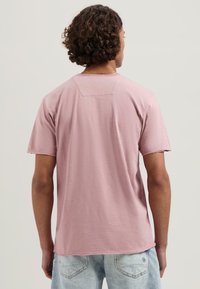 Camisa rosa claro de manga corta con un ajuste relajado, que presenta un pequeño yugo en la parte de atrás y una sutil textura, combinada con unos pantalones cortos de mezclilla azul desgastados.