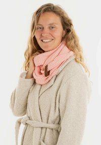 Gehaakte roze infinity sjaal met een gestructureerd patroon, voorzien van een bruine leren label, gedragen over een lichtgrijze donzige badjas.