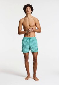 Turquoise zwembroek met een geometrisch wit patroon, elastische tailleband met trekkoord en zijzakken. Model staat op blote voeten.