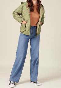 Prošívaná zelená bunda s kapucí, která má kapsy a růžové detaily, kombinovaná s světle modrými širokými džíny a černými teniskami.