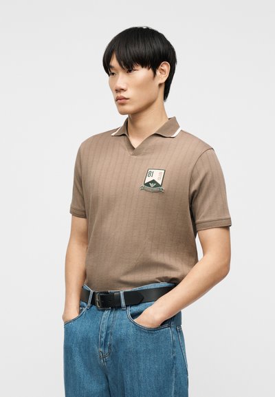 Άνδρας που φοράει καφέ πλεκτό πόλο με κοντά μανίκια και σημαιάκι στο στήθος, μπλε τζιν και μαύρη ζώνη, στέκεται με τα χέρια στις τσέπες.