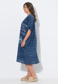 Kaftan bleu marine en tissu léger avec des motifs de vagues blancs et du texte imprimé ; coupe ample et longueur genoux. Porté avec des sandales beiges.