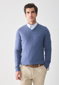 Niebieski sweter w serek z miękkiego materiału, z małym logo na lewej piersi. Noszony na jasnoniebieskiej, w paski koszuli z kołnierzykiem.