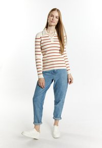 Cremefarbener gerippter Pullover mit roten Streifen, Kragen und drei Knöpfen, kombiniert mit hellblauen hochgekrempelten Jeans und weißen Sneakern.