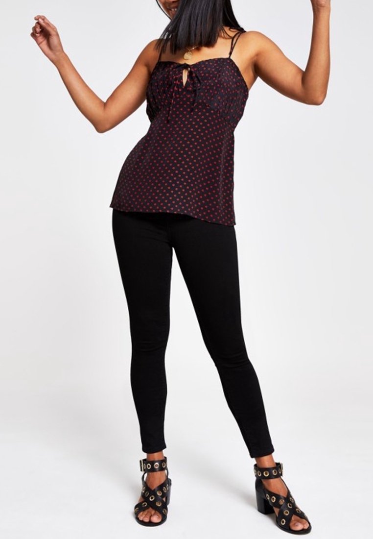 Un débardeur noir et rouge à pois avec un devant noué, des bretelles fines, associé à un legging noir et des sandales à talons hauts noires à brides avec des accents dorés.