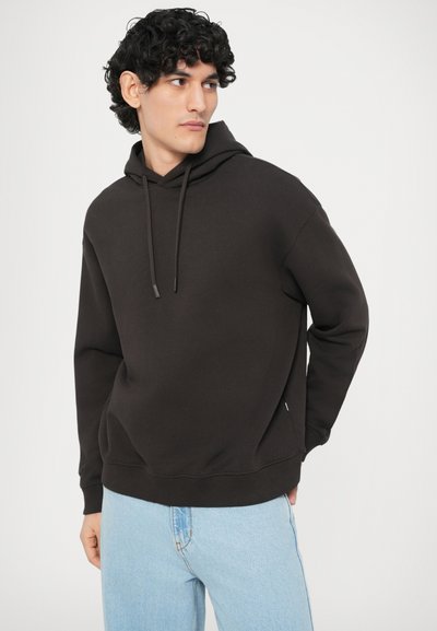 Φούτερ με κουκούλα σε σκούρο καφέ βαμβακερό μείγμα, χαλαρή εφαρμογή με κορδόνια, ριμπ μανσέτες και τελείωμα, συνδυασμένο με ανοιχτό μπλε τζιν.