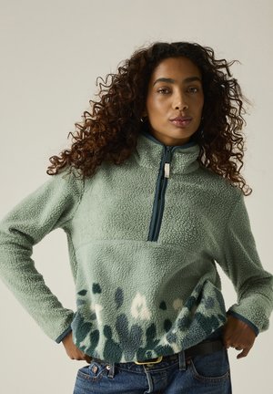 Mujer con cabello rizado que lleva un pulóver polar verde con mitad inferior estampada y cremallera hasta la mitad, posando con las manos en las caderas sobre un fondo liso.
