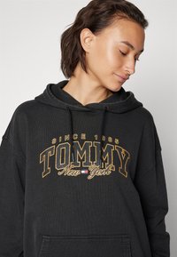 Hanorac negru din bumbac gros, cu o buzunar frontal. Designul include "TOMMY" brodat în aur și un detaliu mic cu steagul.