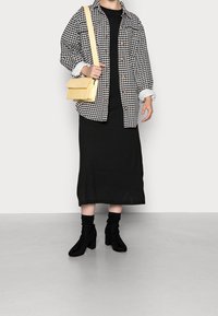 Camicia giacca oversize a scacchi bianco e nero, vestito nero lungo fino al ginocchio, borsa a tracolla gialla e stivali neri con tacco alto e morbidi.