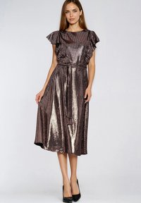 Kobieta ubrana w metaliczną, brązową sukienkę midi z falbanowymi rękawami, paskiem wiązanym w talii i czarne szpilki, stojąca na jednolitym tle.