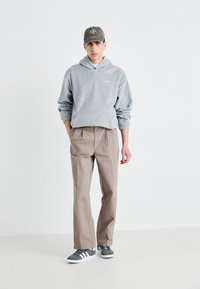 Felpa grigia in pile con tasca a kangaroo, pantaloni leggeri marrone chiaro a gamba affusolata e sneaker grigie con suola bianca. Cappellino nero con logo.