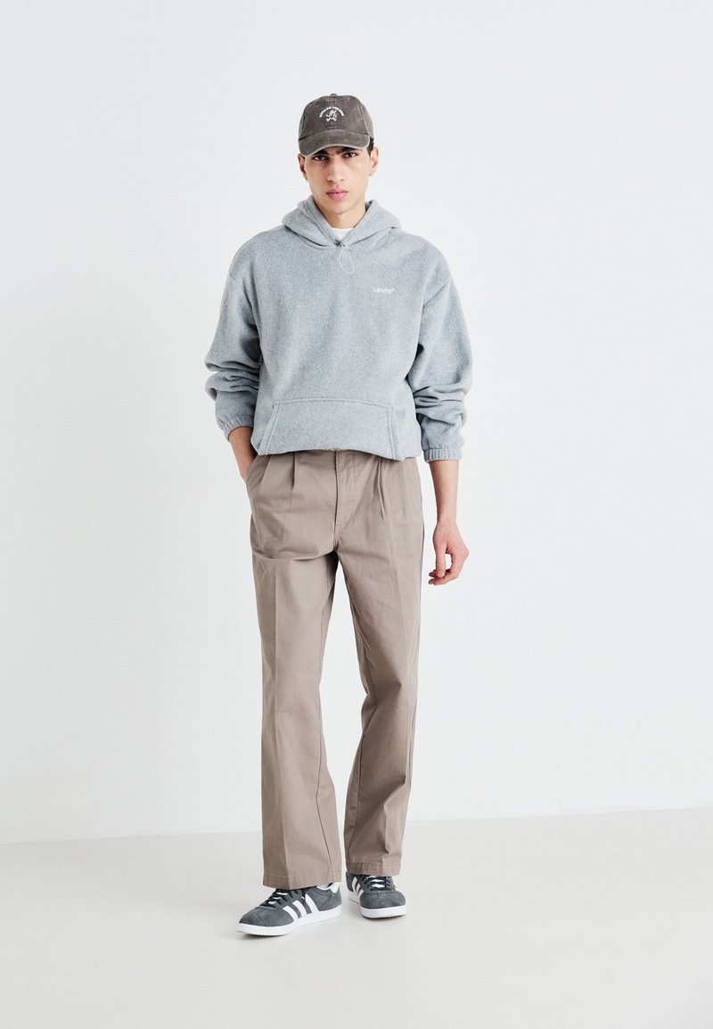 Felpa grigia in pile con tasca a kangaroo, pantaloni leggeri marrone chiaro a gamba affusolata e sneaker grigie con suola bianca. Cappellino nero con logo.
