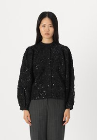 Schwarze Jacke, verziert mit blumigen Pailletten, mit Rundhalsausschnitt und Puffärmeln. Kombiniert mit grauen Nadelstreifenhosen.