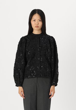 Schwarze Jacke, verziert mit blumigen Pailletten, mit Rundhalsausschnitt und Puffärmeln. Kombiniert mit grauen Nadelstreifenhosen.