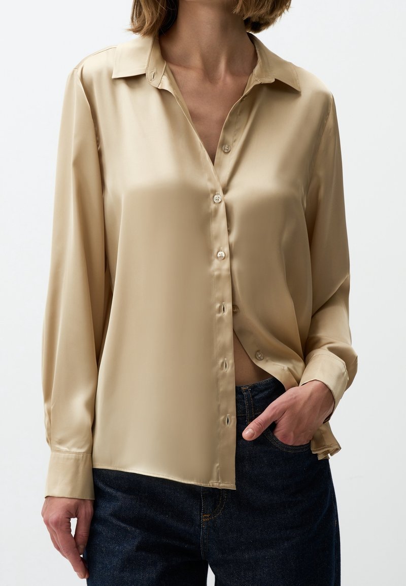 Femme portant une chemise beige satinée brillante à boutons avec manches longues et un jean bleu foncé, une main dans la poche, sur un fond uni.