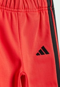 Czerwone sportowe szorty uszyte z miękkiego materiału, z czarną pionową linią i czarnym logo po lewej stronie. Elastyczny pas dla wygody.
