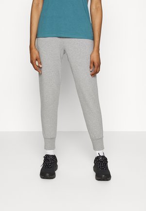 Person iført grå joggingbukser, sorte sneakers med hvide sokker og en teal farvet skjorte, stående mod en ensfarvet hvid baggrund.