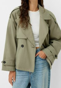 Olivově zelený zkrácený trenčkot s dvojitým zapínáním, velkými klopami a manžetami na knoflíky. V kombinaci s bílým topem a světle modrými džíny.