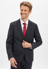 Ciemnoszary garnitur z subtelnym wzorem, klapami w kształcie litery notch i zapięciem na jeden guzik. Połączony z białą koszulą i czerwoną krawatem.