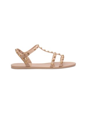 Beige flache Sandale mit drei genieteten Riemen und einem Knöchelriemen mit kleinem goldenen Anhänger, der vom Riemen hängt.