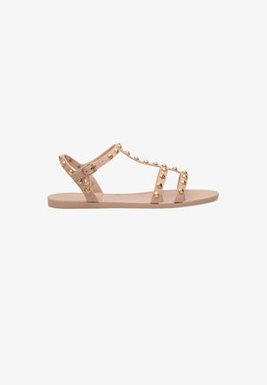 Beige flache Sandale mit drei genieteten Riemen und einem Knöchelriemen mit kleinem goldenen Anhänger, der vom Riemen hängt.