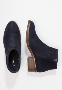 Botines de ante azul marino oscuro con cremallera lateral, puntera redonda y tacón apilado. Detalles de costura en contraste a lo largo de la suela. Textura suave.