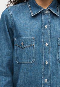 Τζιν πουκάμισο σε μπλε χρώμα με κουμπιά μπροστά, δύο τσέπες στο στήθος και διακοσμητικά καρφιά κατά μήκος του γιακά και των άκρων των τσεπών. Υφή στο ύφασμα.