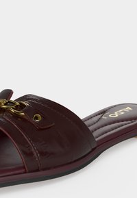 Sandale en cuir marron foncé de type slip-on avec détail chaîne dorée et semelle matelassée portant la marque Aldo, vue de profil sur fond blanc.