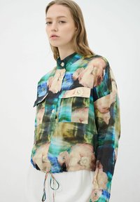 Camisa de estampado floral en tejido ligero con cuello, cierre de botones, dos bolsillos frontales y dobladillo con cuerda ajustable. Los colores incluyen azul, verde y melocotón.