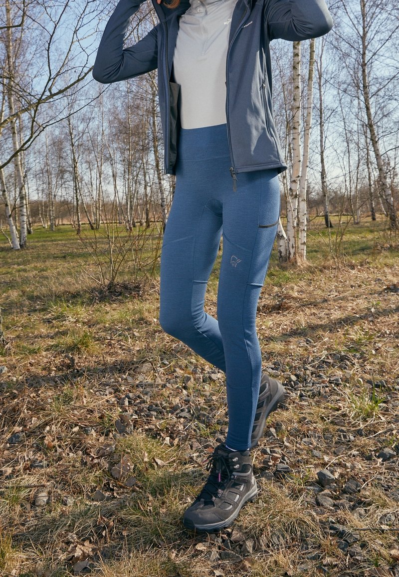 Personne portant des leggings bleus et des chaussures de randonnée noires, debout sur un sol herbeux et rocheux dans une forêt de bouleaux sans feuilles en journée.