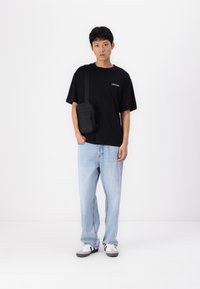 T-shirt noire avec logo blanc, jean bleu clair et baskets grises. Sac bandoulière noir avec fermeture éclair. Fond blanc uni.