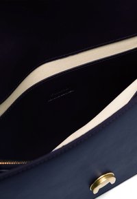 Notranjost modre usnjene torbe s kremno podlogo, opremljena z zlatim okovjem in subtilnim embosiranim logotipom; gladka tekstura.