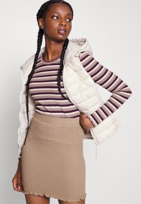 Colete acolchoado em bege claro, com capuz e fecho de zíper. Por baixo, uma blusa ajustada às riscas em roxo e crème com mangas longas. Mini saia texturizada castanha.